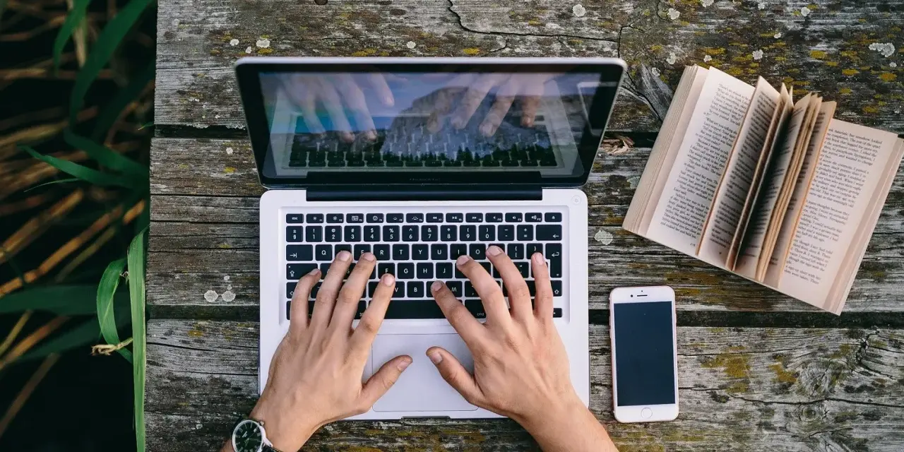Ręce piszą CV funkcjonalne na laptopie, obok leży otwarta książka i telefon.