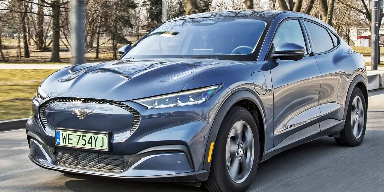 Niebieski Ford Mustang Mach-E jedzie po drodze, z widocznym logo konia na grillu.