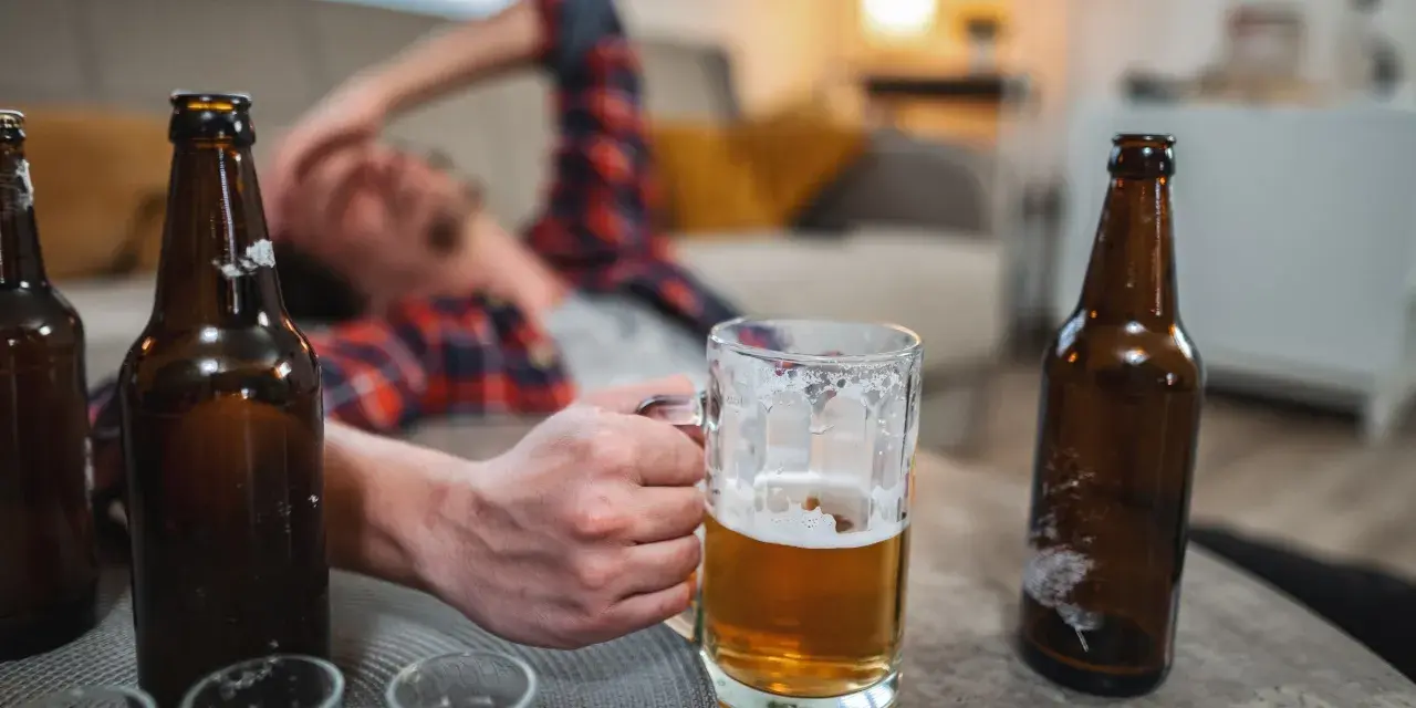Mężczyzna leży na kanapie, trzymając kufel piwa. Wokół puste butelki, sugerujące nagły spadek tolerancji na alkohol.