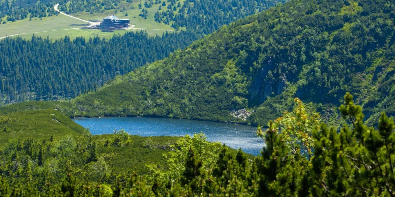 Widok na górskie jezioro i schronisko Samotnia w Karkonoszach. Otoczone zielenią, lasem i skalistymi zboczami.