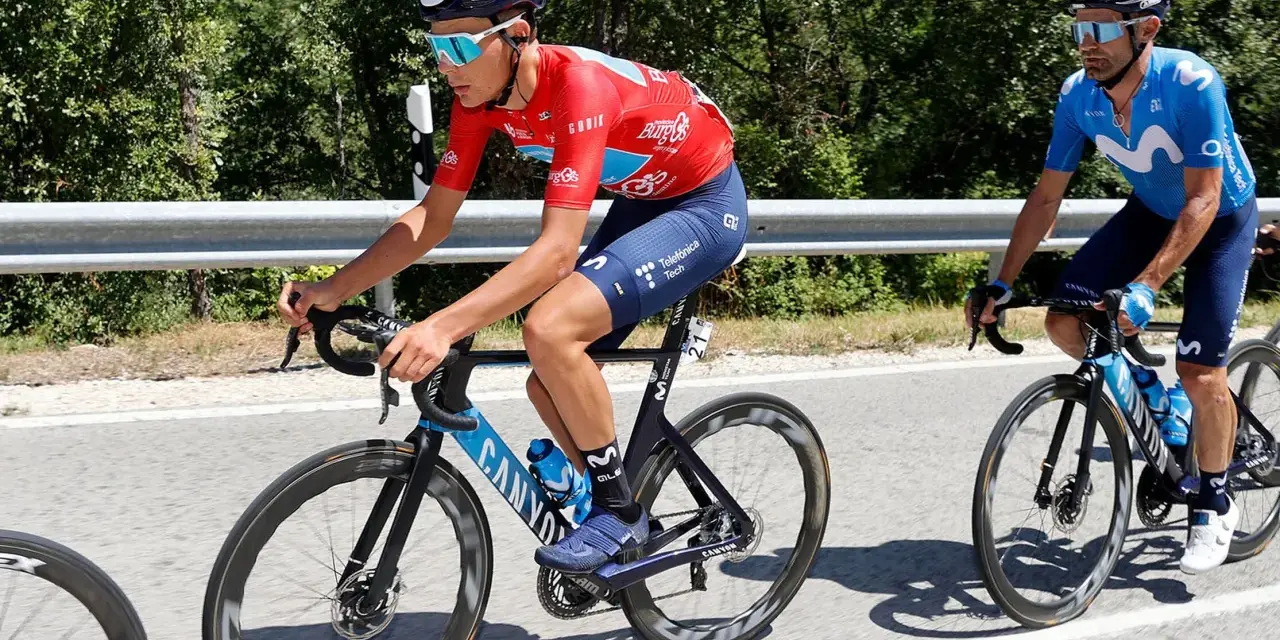 Cyclists in red and blue vuelta jerseys race on a sunny road, with lush green trees lining the background.