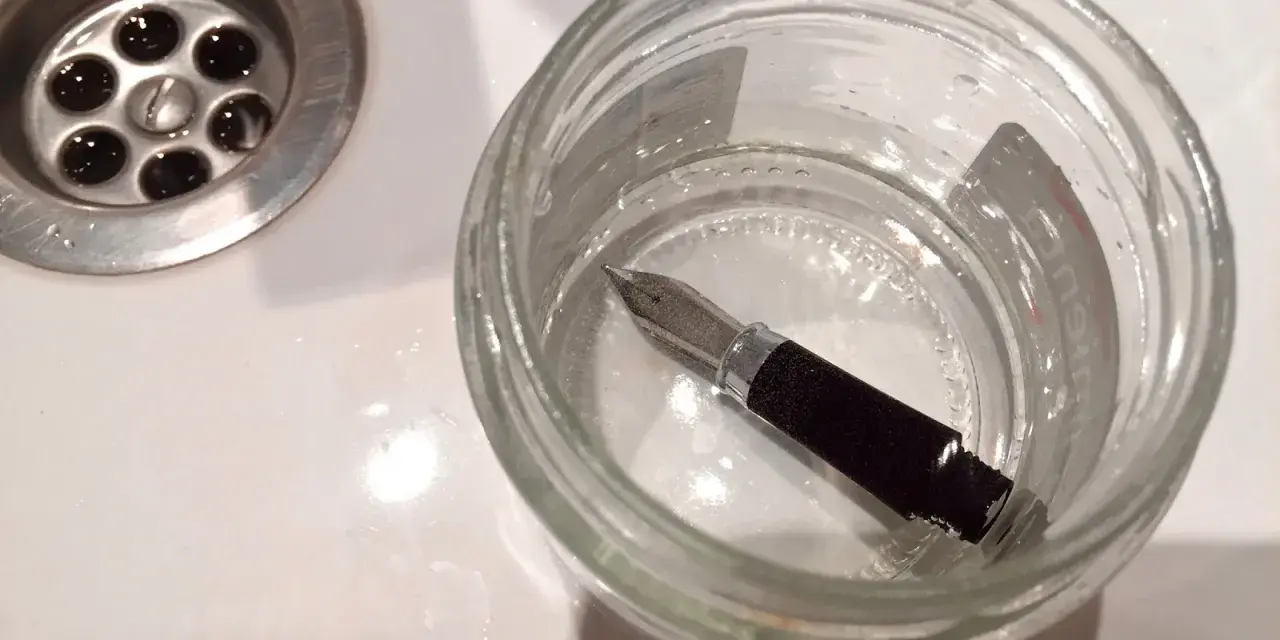 Pluma estilográfica sumergida en un frasco de vidrio con agua, junto a un desagüe. Un paso para aprender como limpiar una pluma.