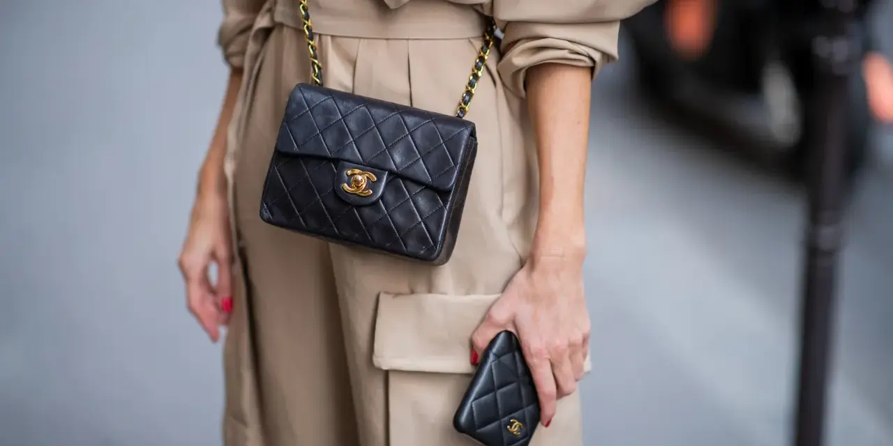 Elegancka kobieta w beżowym płaszczu prezentuje klasyczną, pikowaną torebkę Chanel vintage z złotym łańcuszkiem i logo.