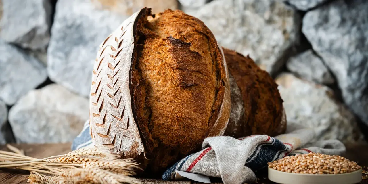 Zwei rustikale Brote, eines mit eingekerbtem Muster, liegen auf einem Holztisch. Weizenähren und eine Schale voller Körner deuten auf ein köstliches Ruchbrot Rezept hin.