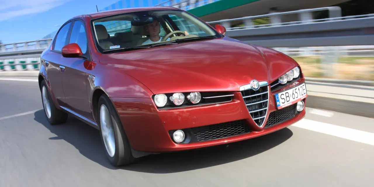Czerwone Alfa Romeo 159 na drodze. Kierowca w białej koszulce, pozytywne opinie o tym modelu potwierdzają jego sportowy charakter.