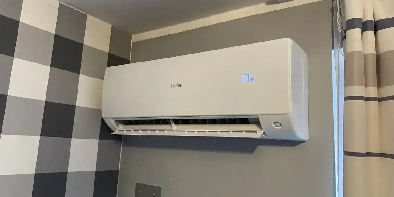Biała klimatyzacja Haier zamontowana na szarej ścianie, obok wzorzystej tapety i zasłon.