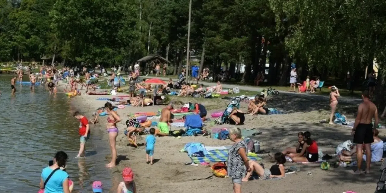 Tłoczna plaża nad jeziorem na Pomorzu. Ludzie opalają się, kąpią i odpoczywają w cieniu drzew.