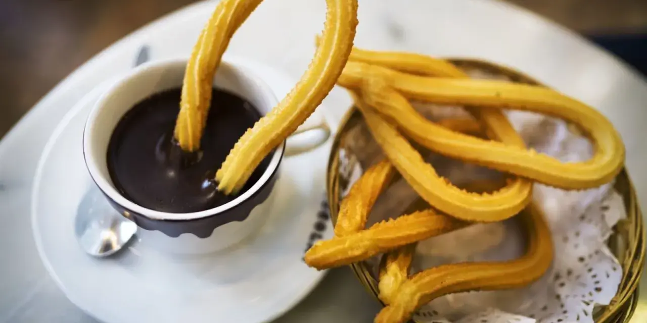 Churros sin gluten dorados y crujientes, uno sumergido en chocolate caliente, otro en una cesta.