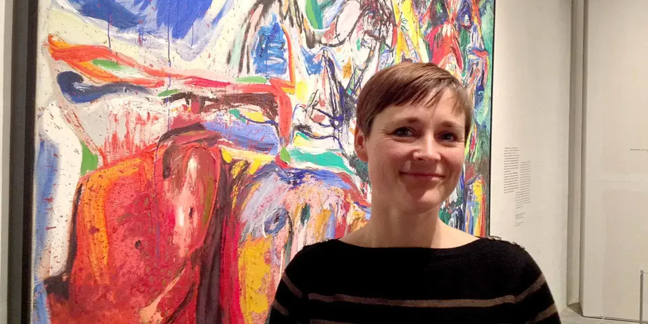 Kvinna ler framför en färgstark, abstrakt målning i Asger Jorns anda.