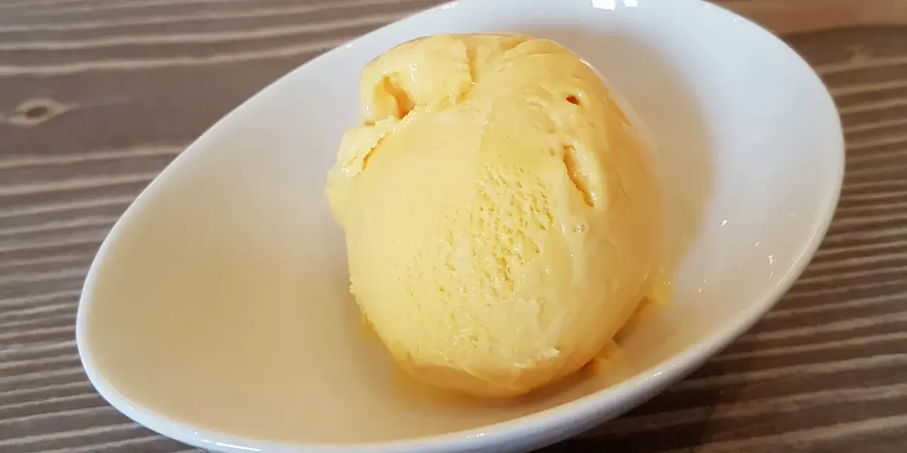 Ein Löffel cremiges, gelbes balla balla Eis in einer weißen Schale auf Holzhintergrund.