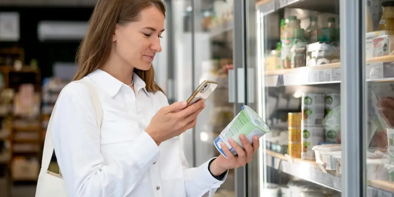 Kobieta w białej koszuli trzyma jogurt i telefon, analizując produkt w sklepie.
