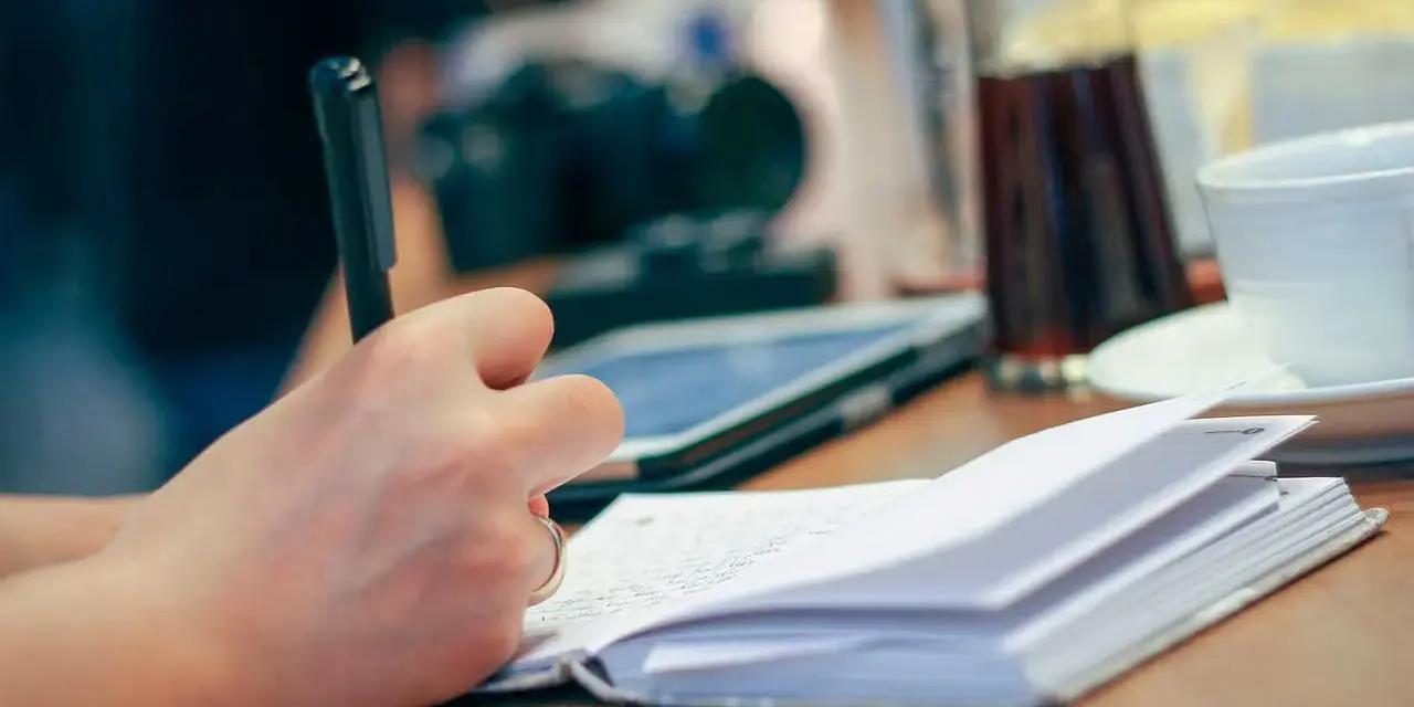 Mano escribiendo en un cuaderno abierto, inspirada para escribir cuentos. En la mesa hay una tableta, una taza y una jarra.
