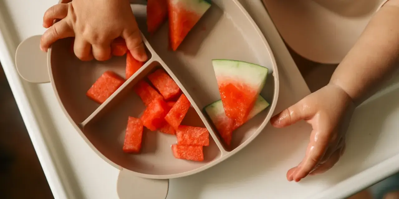Kleine Hände greifen nach saftigen Wassermelone-Stücken auf einem Teller. Ein süßer Wassermelone-Baby-Moment.