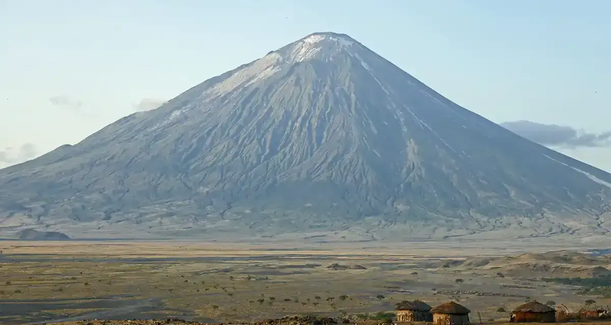 Ol Doinyo Lengai, wulkan w Tanzanii, góruje nad sawanną z kilkoma tradycyjnymi chatami Masajów.
