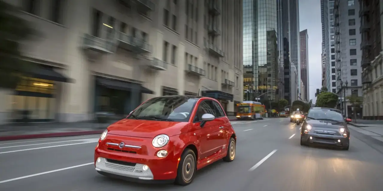 Czerwony Fiat 500e jedzie ulicą w mieście, w tle wieżowce i autobus.