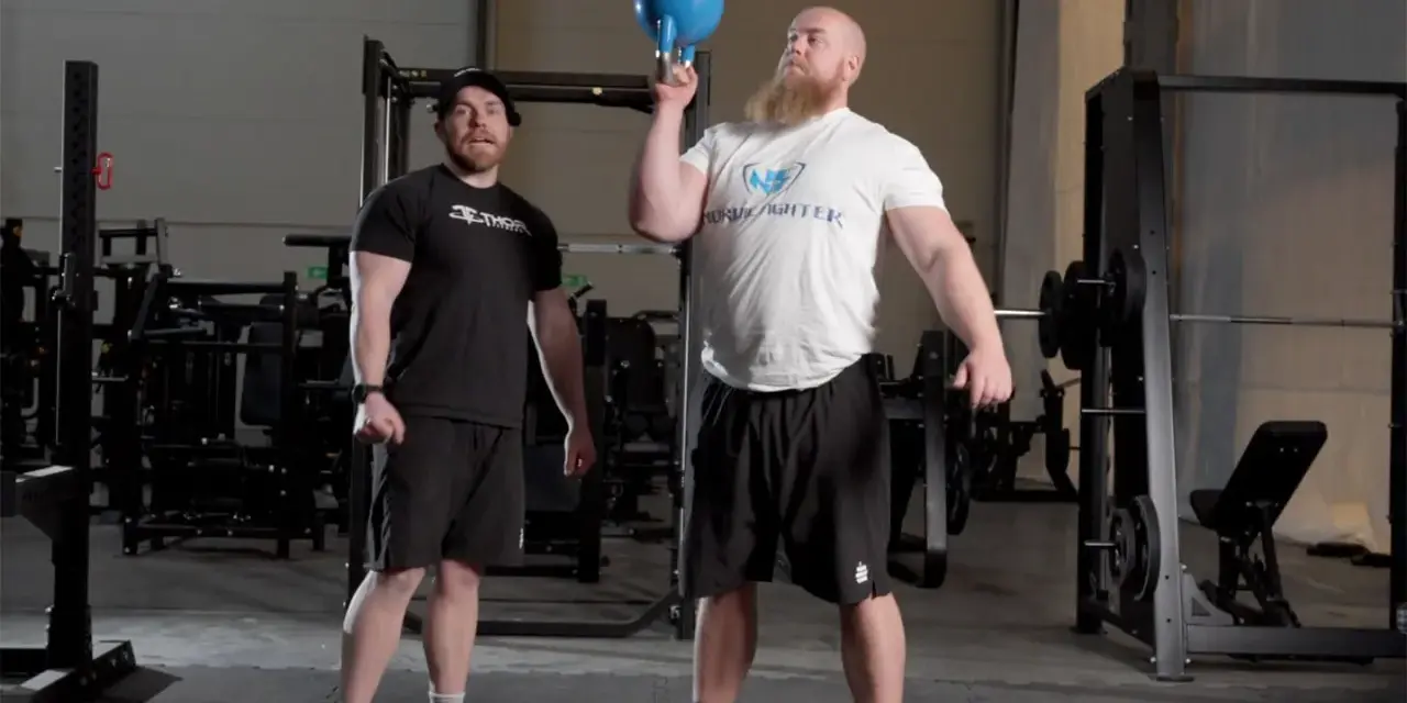 Man med skägg utför en kettlebell press med en blå kettlebell. En annan man står bredvid och tittar.