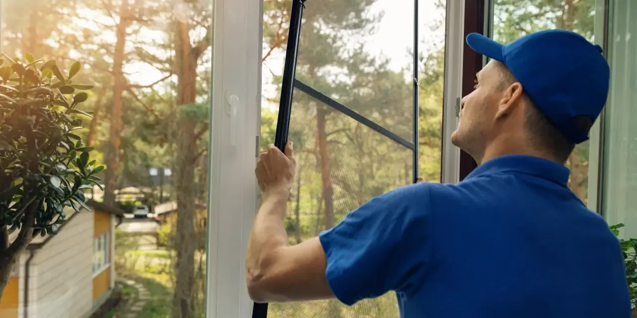 Man in blue shirt and cap performs window screen installation, preparing to fit the mesh into the frame.