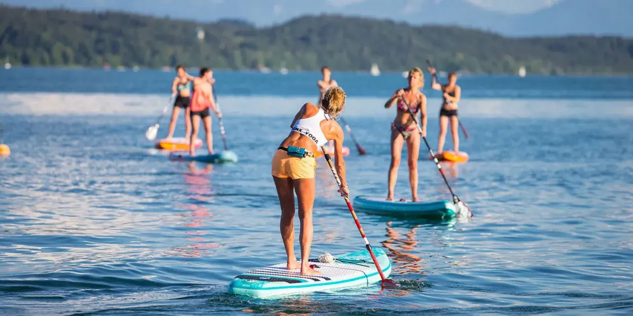 Eine Gruppe von Frauen genießt das Stand-Up-Paddling auf einem See. Sie stehen auf ihren Boards und paddeln durch das ruhige Wasser, während im Hintergrund Berge und Bäume zu sehen sind.