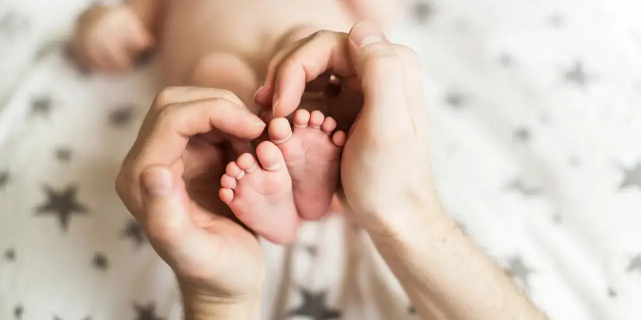 Dłonie obejmujące stópki 7 tygodniowego dziecka. Delikatne paluszki i pięty maluszka otoczone troską.