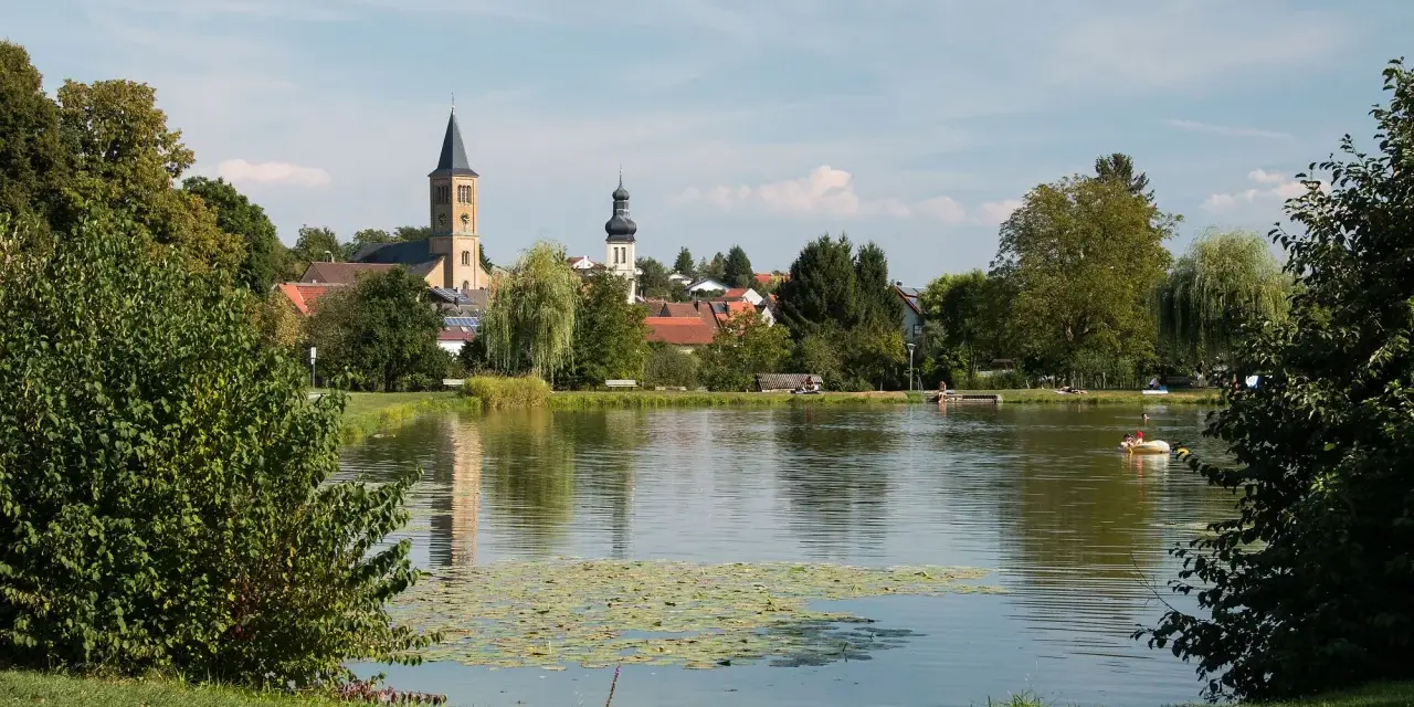Idyllischer Elsenzer See mit Seerosen, Bäumen und einer Kirche im Hintergrund. Ein Boot mit zwei Personen treibt auf dem Wasser.