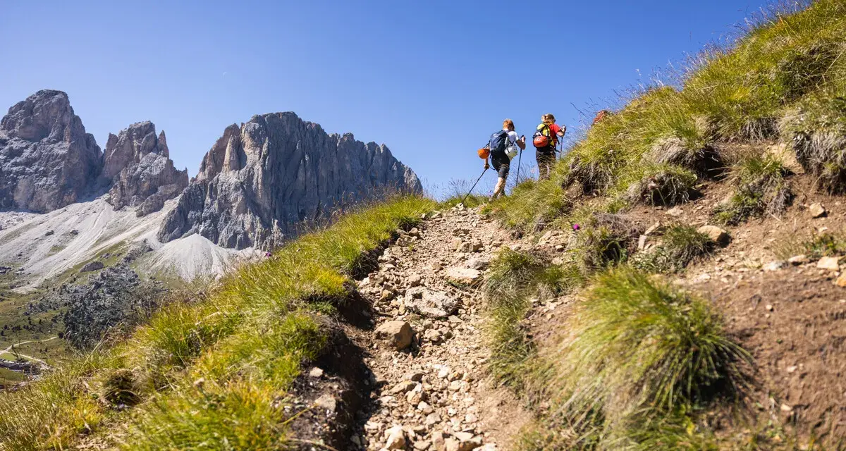 Due escursionisti affrontano un sentiero roccioso sui **cammini in Trentino**, con imponenti vette sullo sfondo.