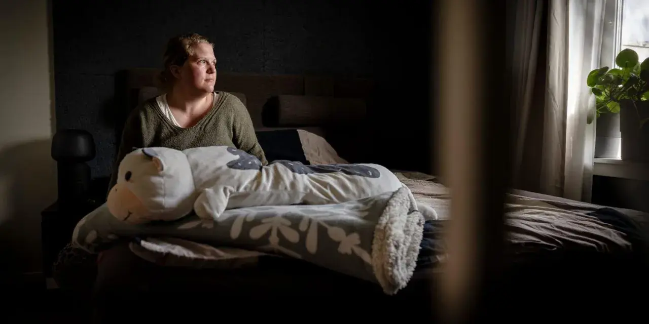 En kvinna sitter i sängen med en stor kramdjursko. Hon ser ut att fundera, kanske över sina två personligheter.