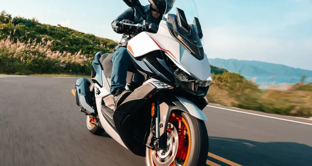 Kymco Xciting 400i dane techniczne: skuter z kierowcą na krętej drodze, z widokiem na góry i jezioro w tle.