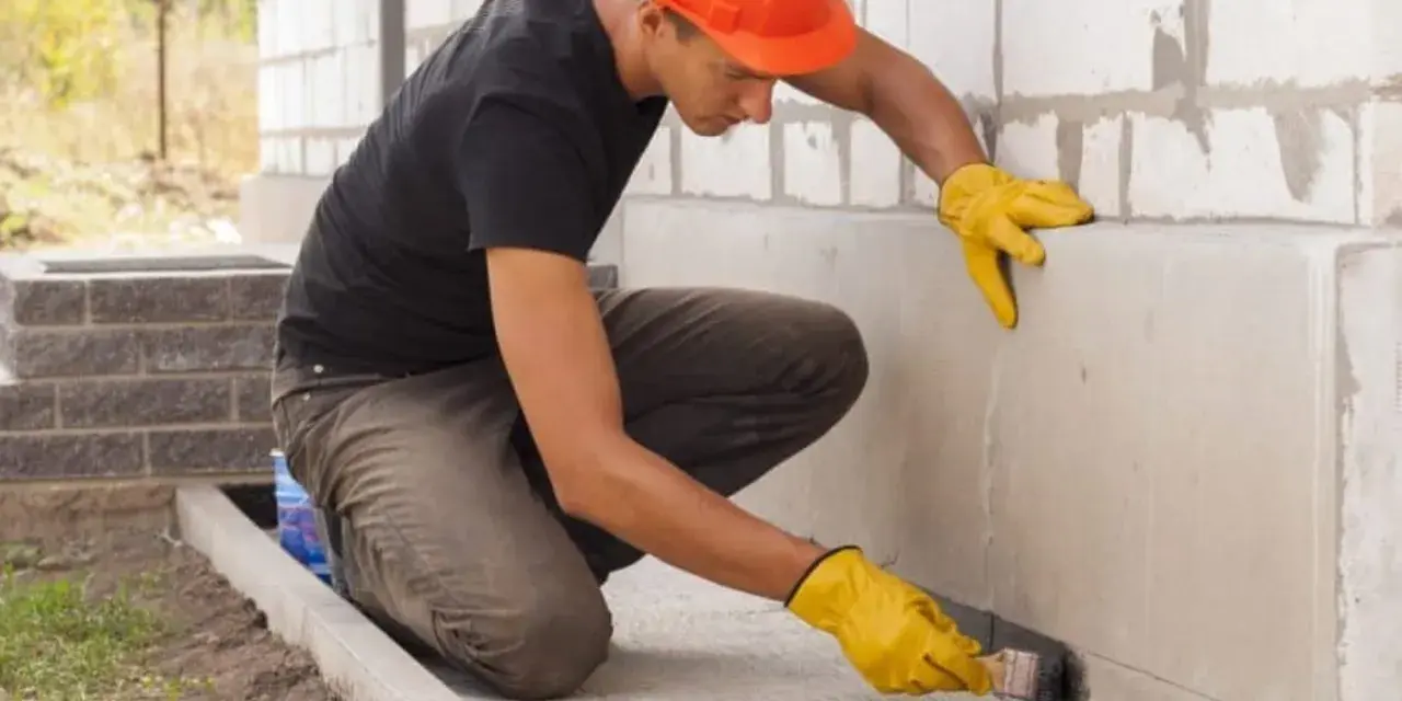 Obrero con casco naranja aplica producto para impermeabilizar pared exterior de bloque de hormigón.