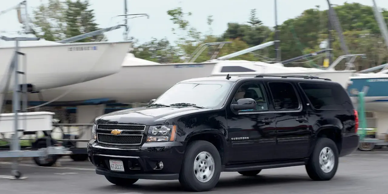 Czarny Chevrolet Suburban jedzie obok przyczep z łodziami, w tle drzewa.