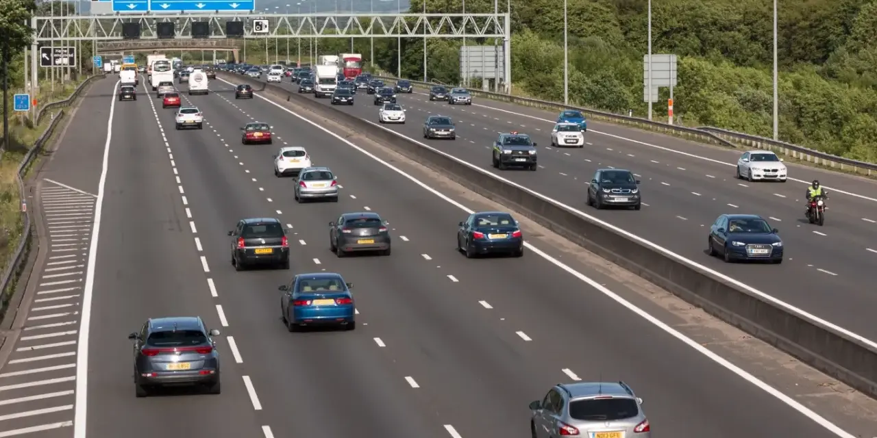 Autostrada z ruchem lewostronnym, pełna samochodów i ciężarówek. Znaki drogowe wskazują M1, A405 i M25.