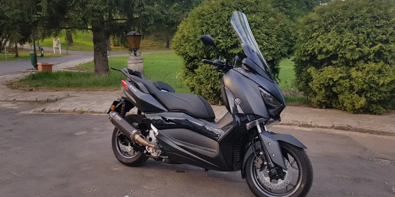 Czarny skuter Yamaha XMAX 300 zaparkowany na tle zieleni i drzew.