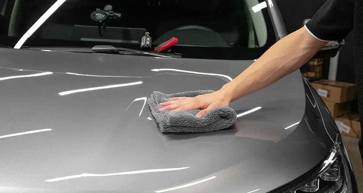 A hand buffs a car's hood with a microfiber cloth, applying a ceramic wax for a deep shine.