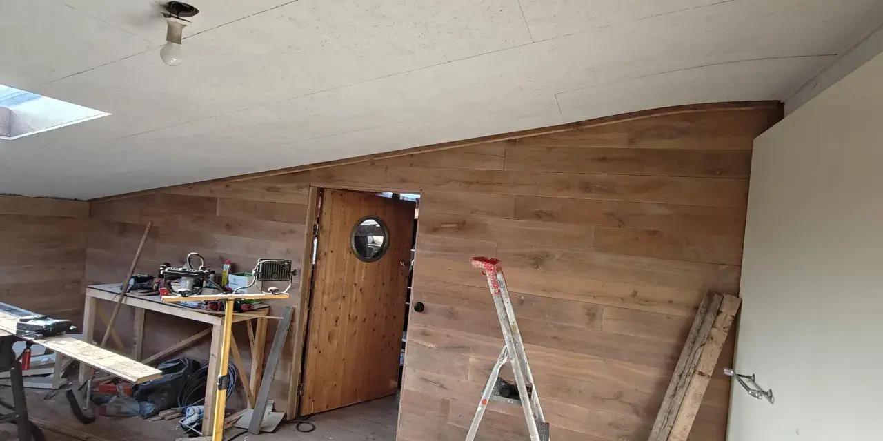 Taket är målat OSB. Rummet är under renovering med träpanel på väggarna, en dörr med runt fönster och byggmaterial utspritt.
