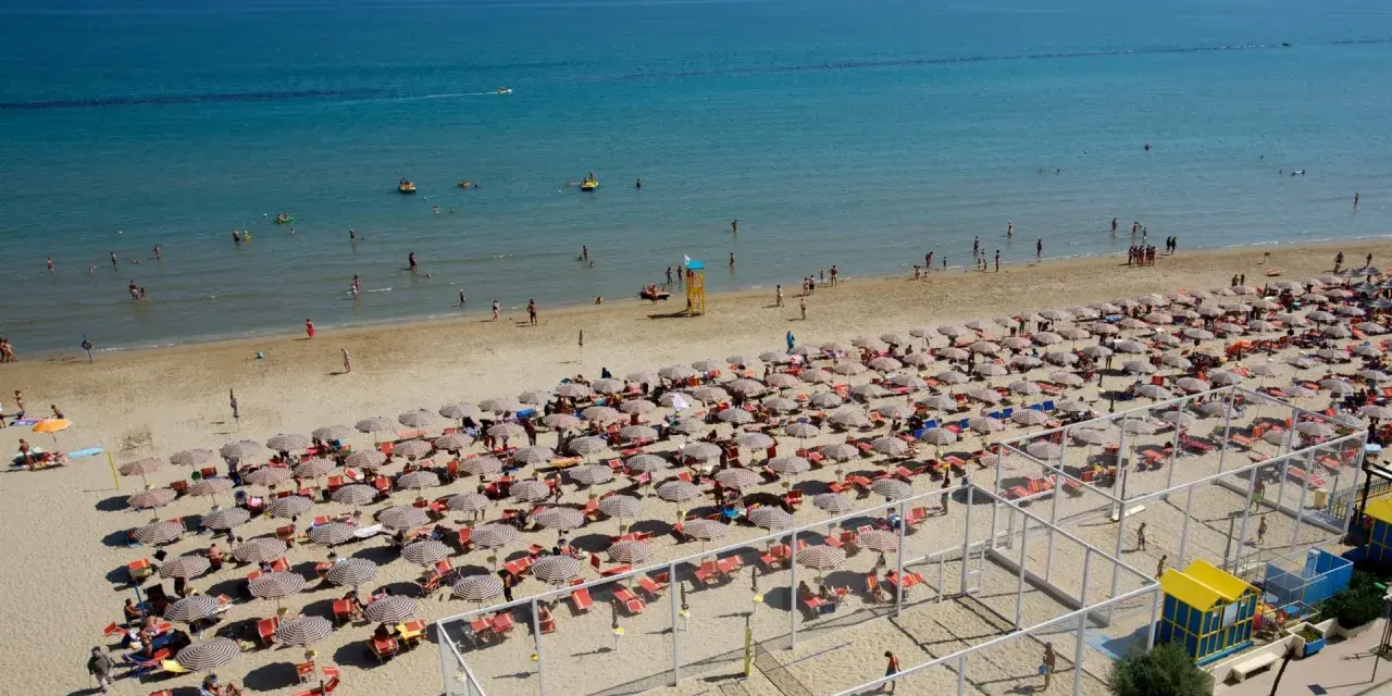 A crowded beach with rows of umbrellas and sunbathers, and people swimming in the blue sea.