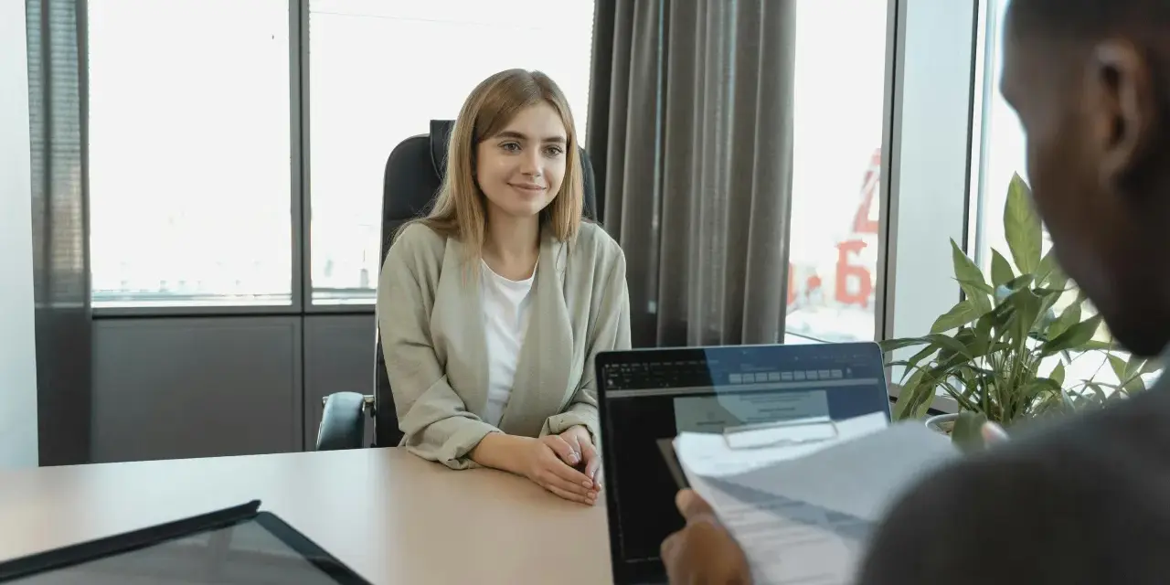Mujer joven sonríe sentada en una oficina, lista para una entrevista. Su atuendo es profesional, demostrando qué usar para una entrevista.