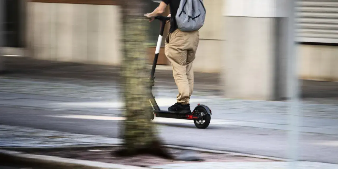 En person på elsparkcykel, med ryggsäck, susar fram på trottoaren. Frågan om får man åka två på elsparkcykel är relevant här.