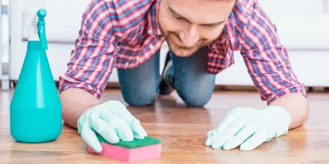 Un homme, muni de gants et d'une éponge, s'agenouille pour nettoyer une tache sur le parquet.