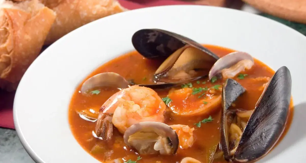 Sopa de mariscos en plato blanco con mejillones, camarones, almejas y pan.