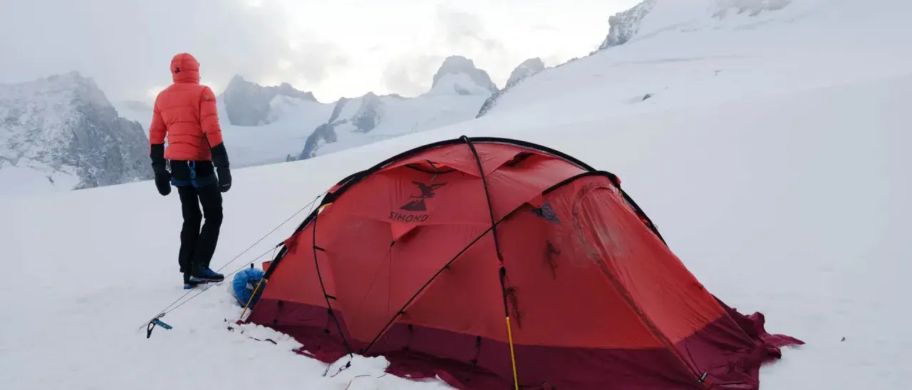 Alpinista w czerwonej kurtce stoi obok namiotu na tle ośnieżonych szczytów. W oddali widać groźne góry, gdzie mogą wystąpić lawiny w Alpach.
