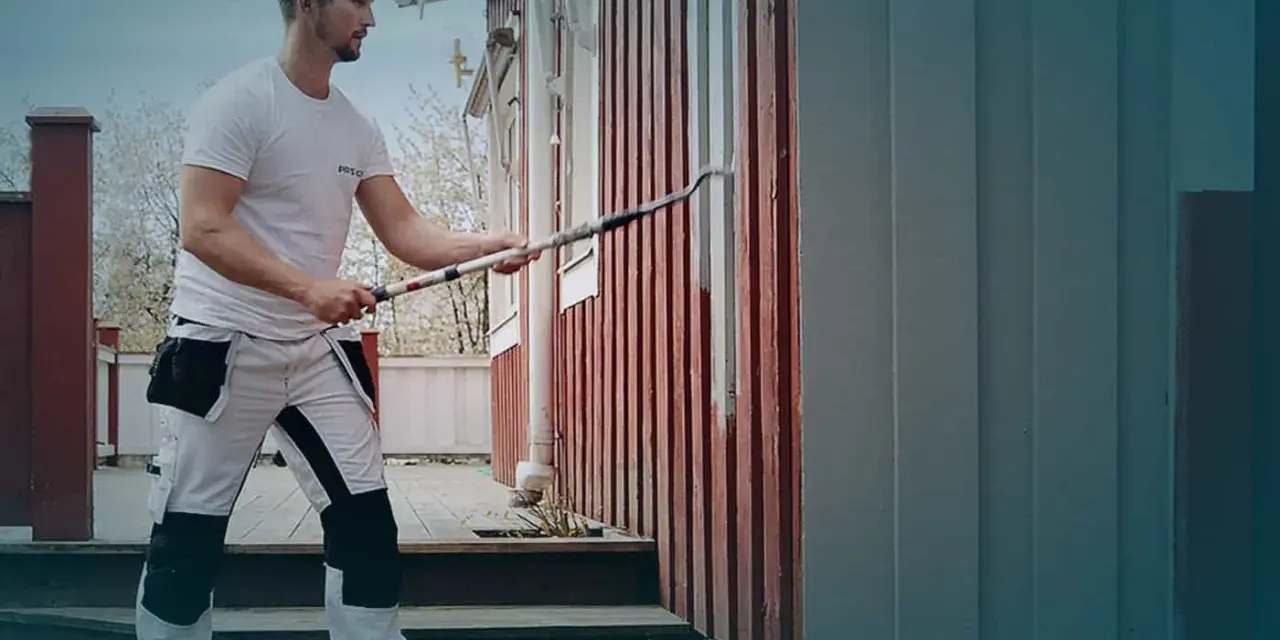 Man i arbetskläder använder en lång roller för att måla huset. En hink med färg står på gräset.