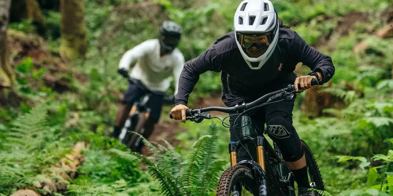 Riders in helmets and protective gear shredding a forest trail. This image shows what to wear mountain biking: full-face helmets, goggles, long sleeves, and knee pads.