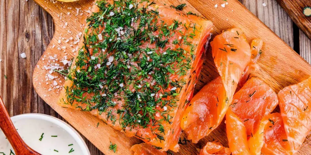 En bit gravad lax, generöst toppad med dill och flingsalt, ligger på en träskärbräda bredvid skivor av lax och en skål med sås.