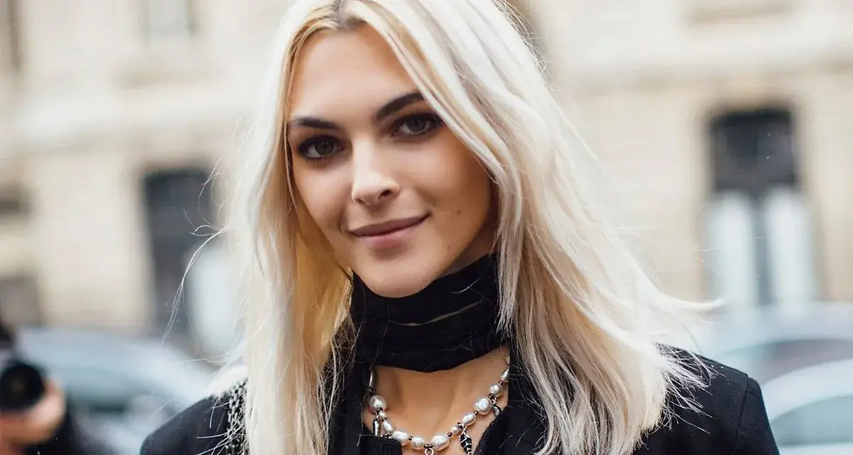 Kobieta z platynowym blondem, w czarnym golfie i naszyjnikiem z pereł, uśmiecha się lekko.