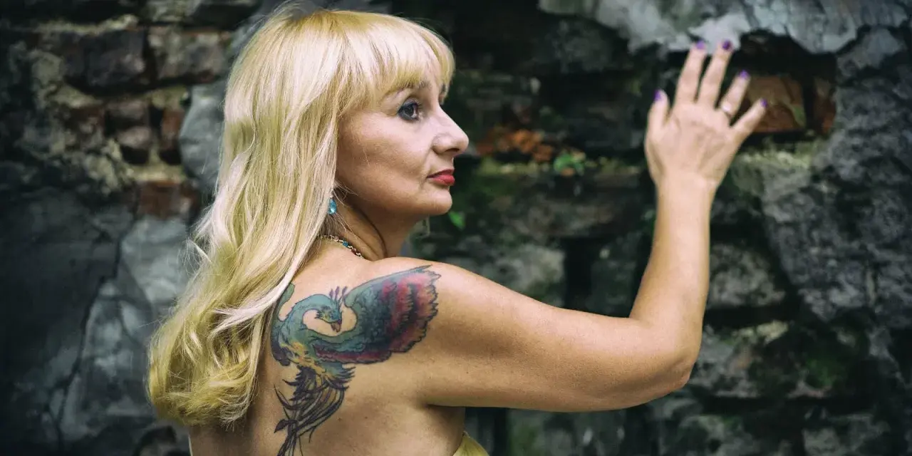 Blondynka z tatuażem feniksa na plecach opiera się o kamienną ścianę. Czyżby moda na tatuaże mijała, a ona wciąż pielęgnuje swoje?