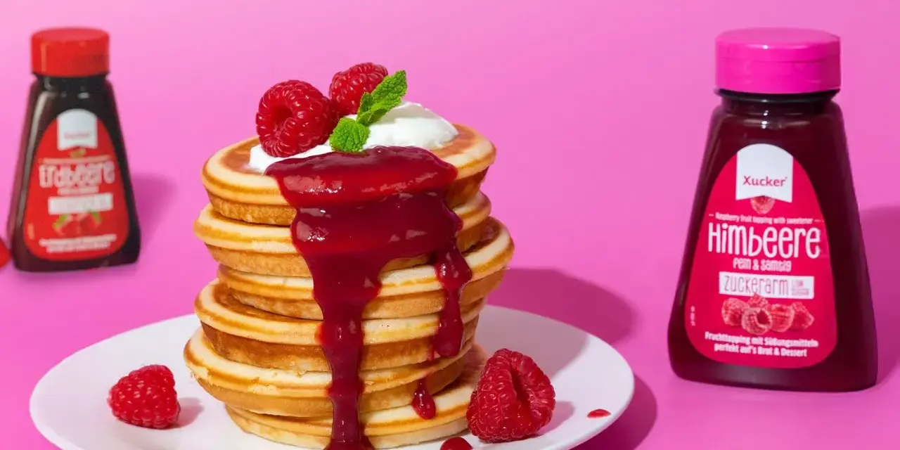 Ein Stapel zuckerfreie pancakes mit Himbeersauce und frischen Beeren. Daneben Flaschen mit Erdbeer- und Himbeer-Topping.
