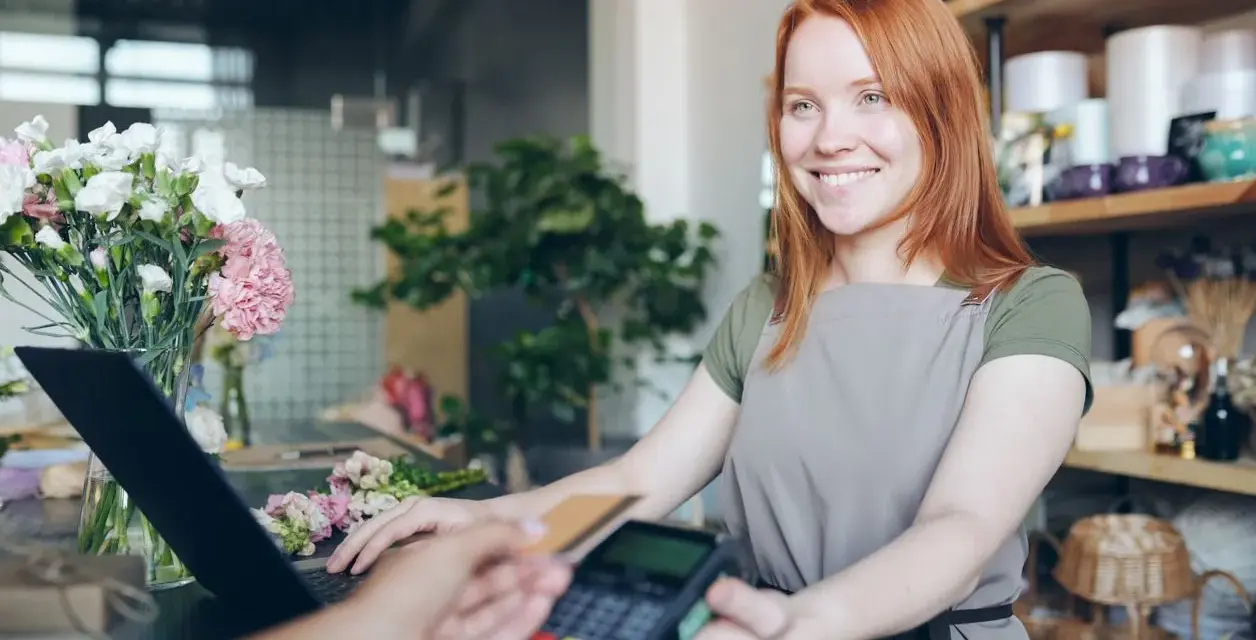 Uśmiechnięta sprzedawczyni w kwiaciarni obsługuje płatność kartą. Na ladzie leży laptop i bukiet kwiatów. Klient płaci zbliżeniowo, a sprzedawczyni przygotowuje paragon z nipem.