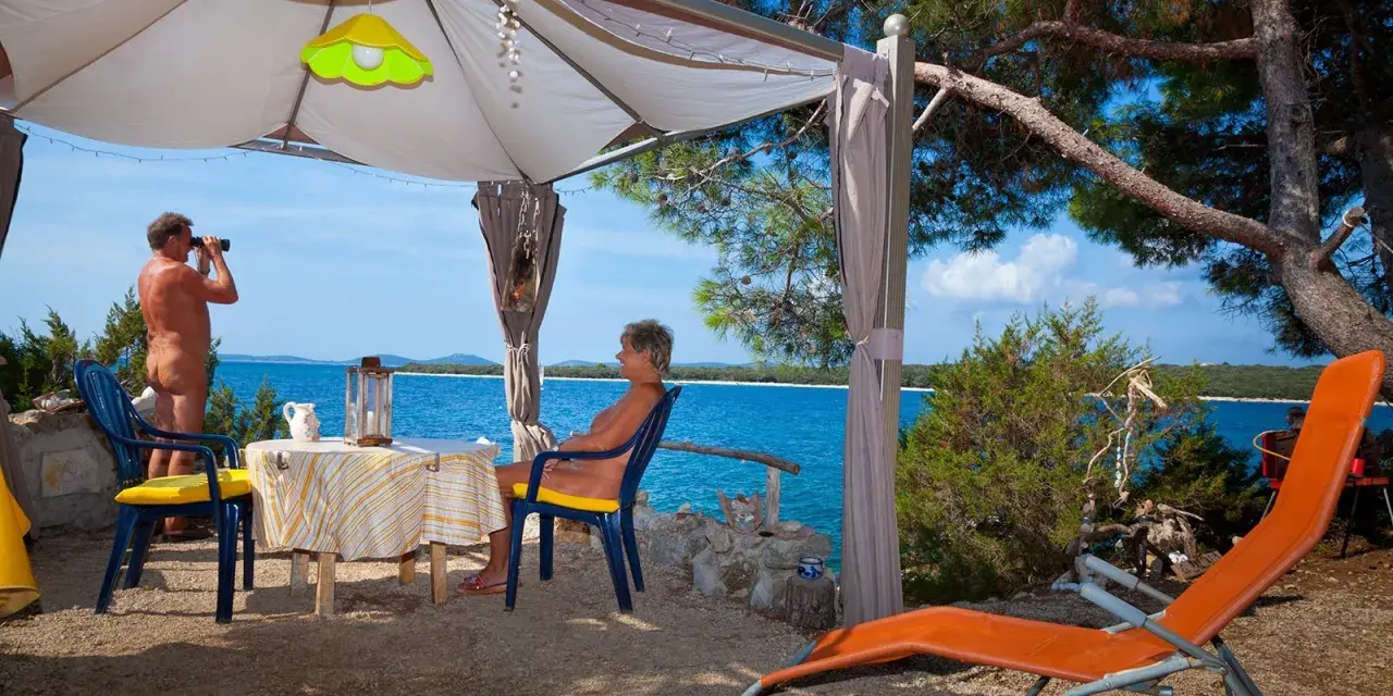 Para na plaży, relaksująca się w namiocie. Mężczyzna obserwuje morze przez lornetkę. To idealne miejsce na naturystyczny camping.