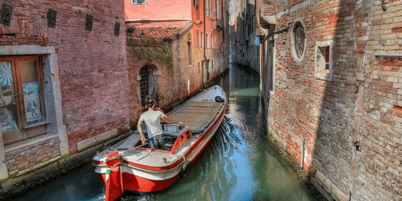 Un uomo guida una barca su un canale stretto a Venezia, circondato da edifici in mattoni. Quanti canali ci sono a Venezia? Molti, come questo!