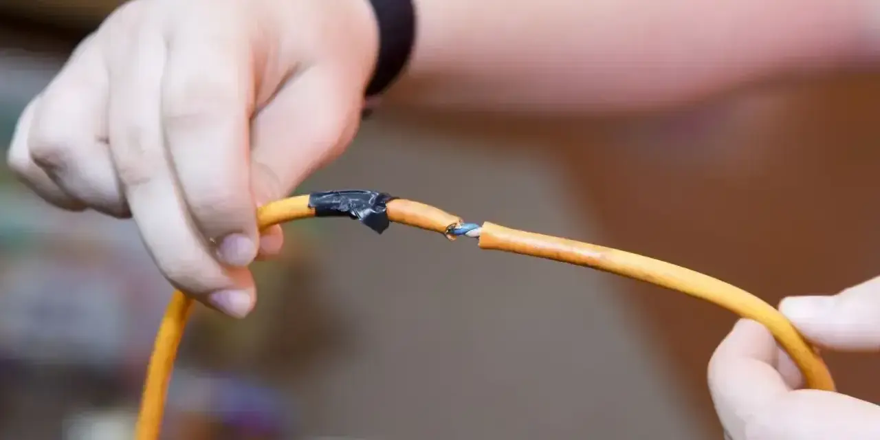 Dłonie trzymające pomarańczowy kabel z odsłoniętymi żyłkami, prowizorycznie połączone taśmą. Pokazuje, jak połączyć kable bez lutowania.