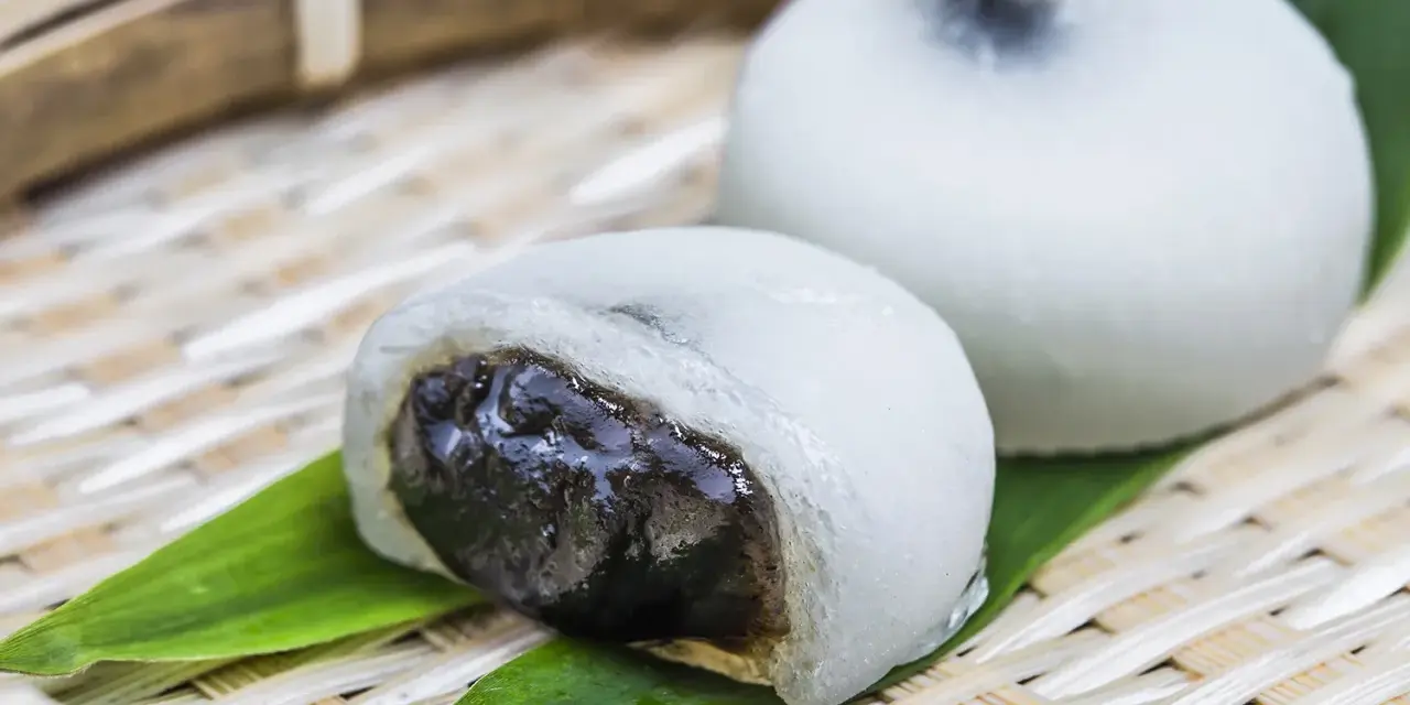 Dwa białe mochi, jedno przekrojone, ukazujące ciemne nadzienie. Podane na bambusowej tacce z liśćmi.